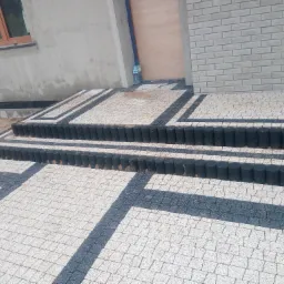 Nowo ułożona kostka brukowa w szaro-białej kolorystyce, tworząca geometryczny wzór przed wejściem do domu z szarą elewacją i drewnianym oknem, z czarnymi obrzeżami schodów i dwiema butelkami wody...