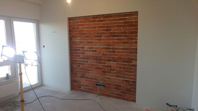 Ściana z cegły dekoracyjnej w trakcie montażu, widoczne gniazdka elektryczne, lampa budowlana, surowy stan wykończenia pomieszczenia.