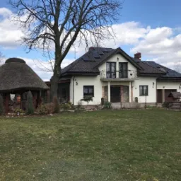 Dom jednorodzinny z panelami słonecznymi na dachu, studnią i altaną krytą strzechą w ogrodzie.