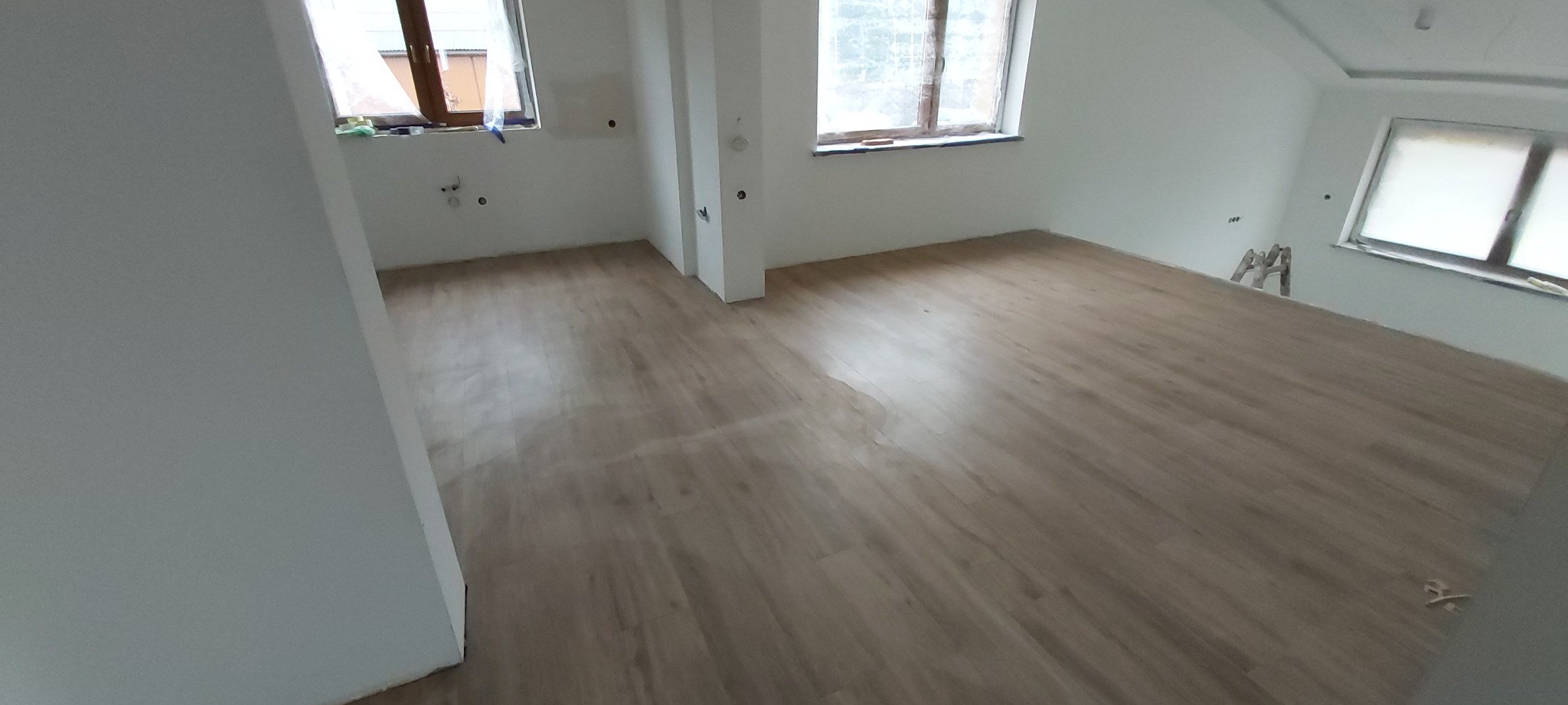 Jasna podłoga z paneli imitujących drewno w nowo wykończonym wnętrzu z trzema oknami, ściany pomalowane na biało, widoczne ślady po instalacjach elektrycznych, drabina oparta o ścianę.