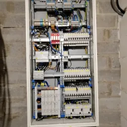 Otwarta skrzynka elektryczna z bezpiecznikami, zabezpieczeniami przepięciowymi i przewodami, zamontowana na ścianie z szarego betonu komórkowego.