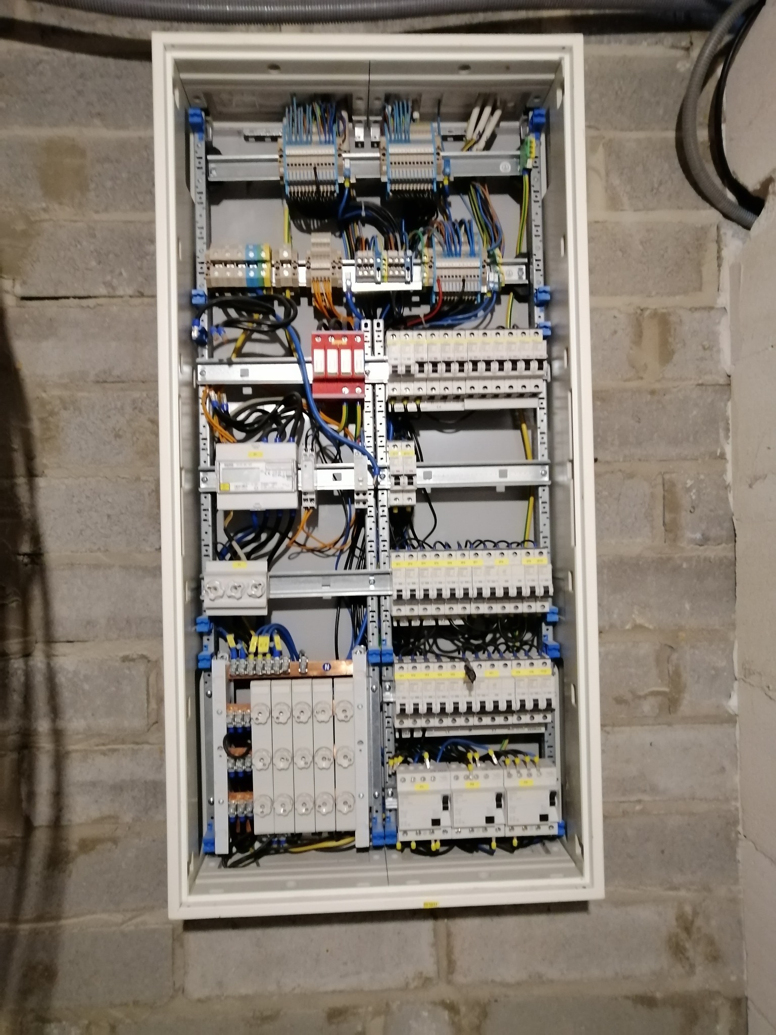 Otwarta skrzynka elektryczna z bezpiecznikami, zabezpieczeniami przepięciowymi i przewodami, zamontowana na ścianie z szarego betonu komórkowego.