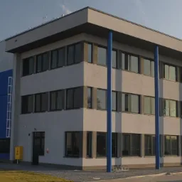 Nowoczesny budynek magazynowy z rampami załadunkowymi, niebiesko-szary, z logo firmy na fasadzie, parking z kilkoma samochodami z przodu.