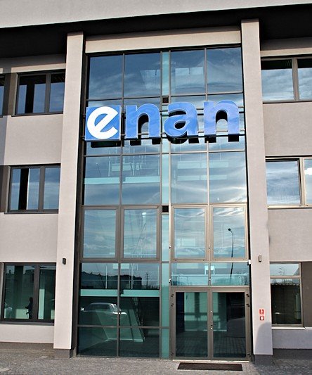 Elewacja budynku biurowego z logo firmy 'enan' nad wejściem, widoczne odbicie samochodu w szklanej fasadzie.