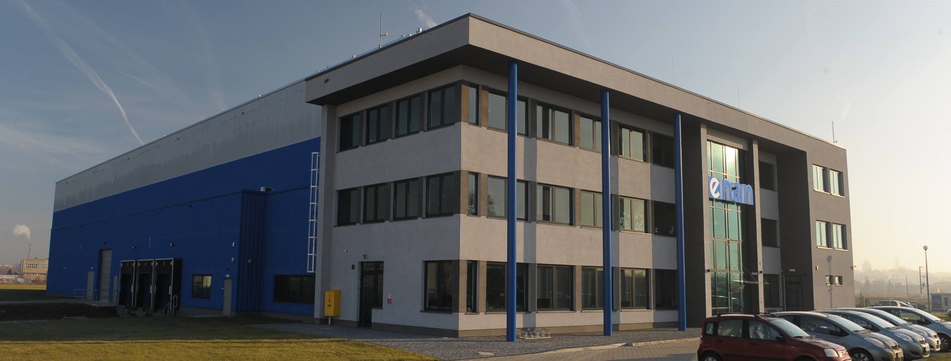 Nowoczesny budynek magazynowy z rampami załadunkowymi, niebiesko-szary, z logo firmy na fasadzie, parking z kilkoma samochodami z przodu.