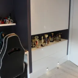 Pok&oacute;j młodzieżowy z biurkiem, krzesłem gamingowym i białymi meblami na wymiar, wnęka z trofeami i nagrodami.