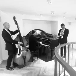 Czarno-białe zdjęcie zespołu muzycznego podczas występu: kontrabasista z uśmiechem, pianista przy fortepianie z dekoracyjnym oknem w tle i trębacz w okularach, wszyscy w smokingach, na parkiecie...