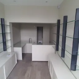 Nowoczesne wnętrze sklepu lub biura z minimalistycznymi szklanymi półkami na ciemnych wspornikach, białymi szafkami i ladą, widok w głąb pomieszczenia z drzwiami.