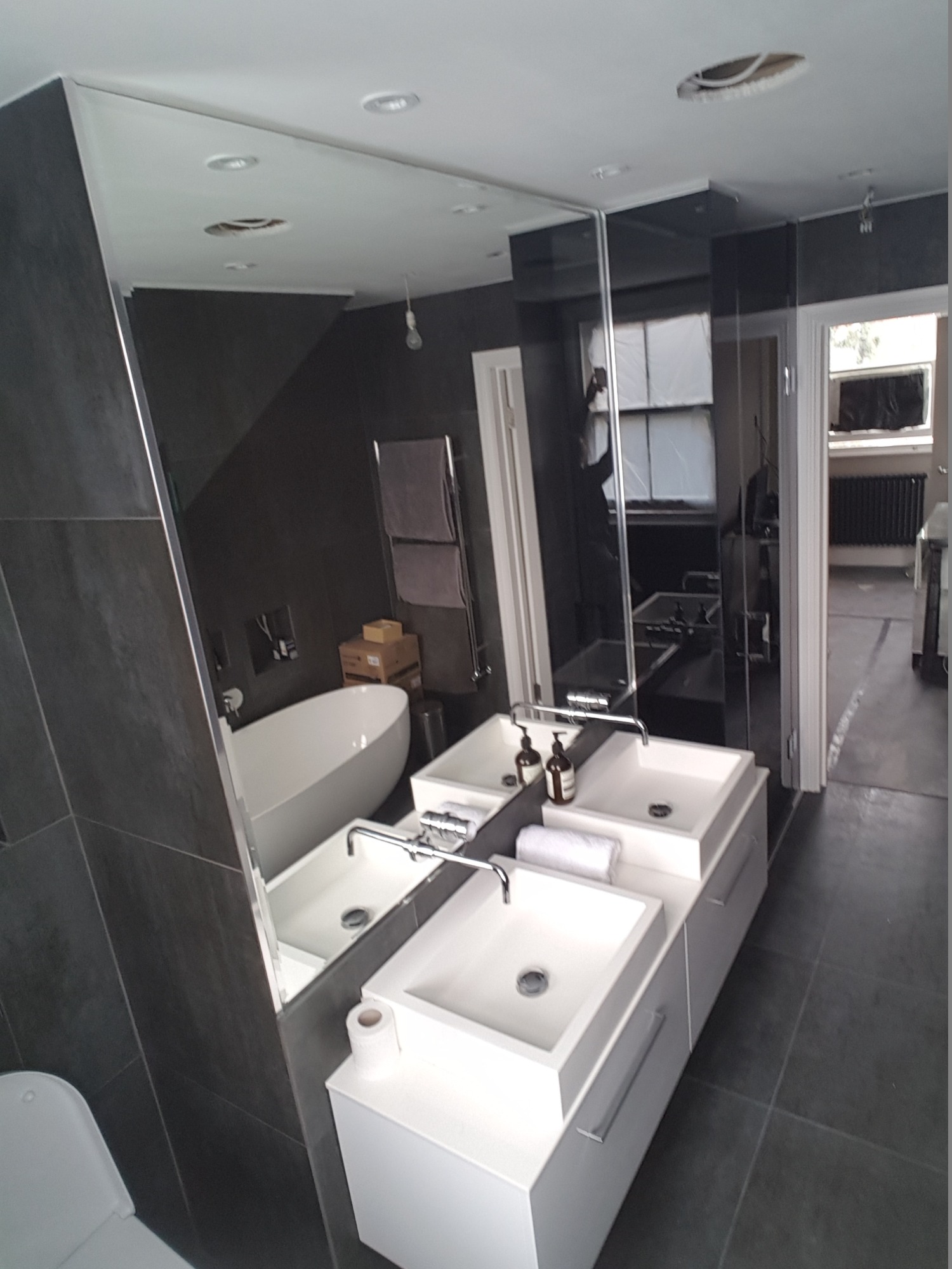 Nowoczesna łazienka z dwoma umywalkami wpuszczonymi w białe szafki, duże lustro bez ramy na ścianie z ciemnych płytek, widoczna wanna i grzejnik drabinkowy.