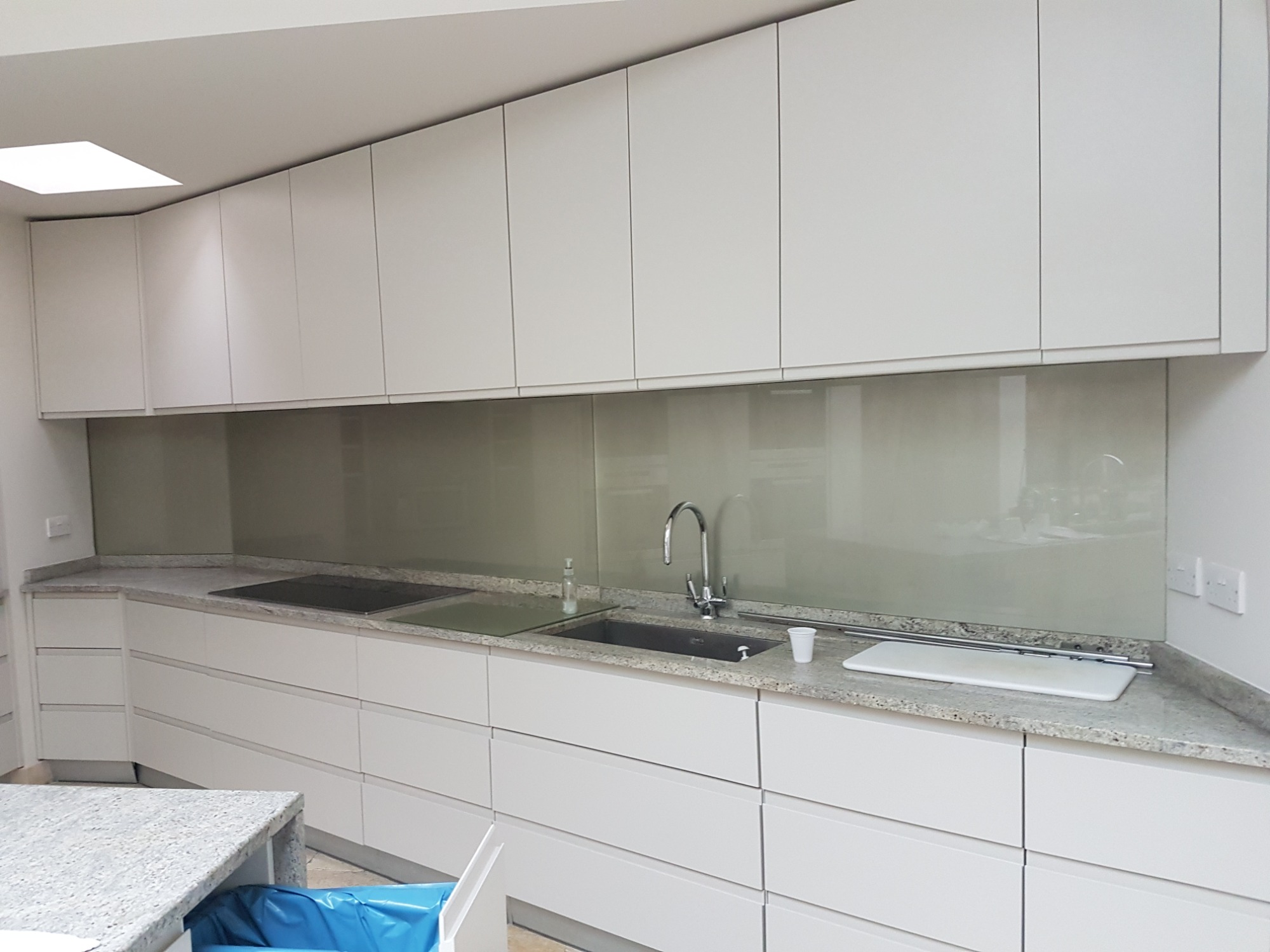 Nowoczesna kuchnia z białymi szafkami, blatem z szarego granitu i szklanym panelem ściennym w odcieniach szarości, minimalistyczny design.