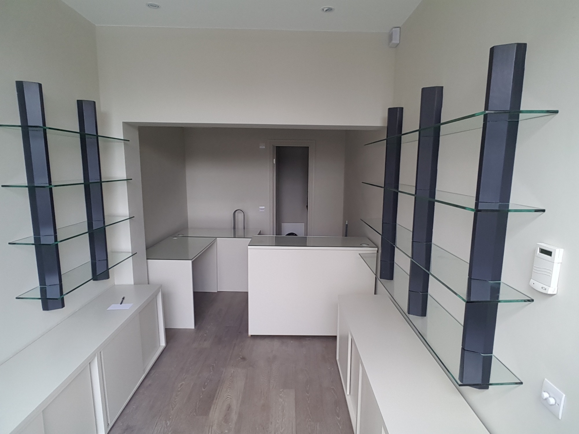 Nowoczesne wnętrze sklepu lub biura z minimalistycznymi szklanymi półkami na ciemnych wspornikach, białymi szafkami i ladą, widok w głąb pomieszczenia z drzwiami.