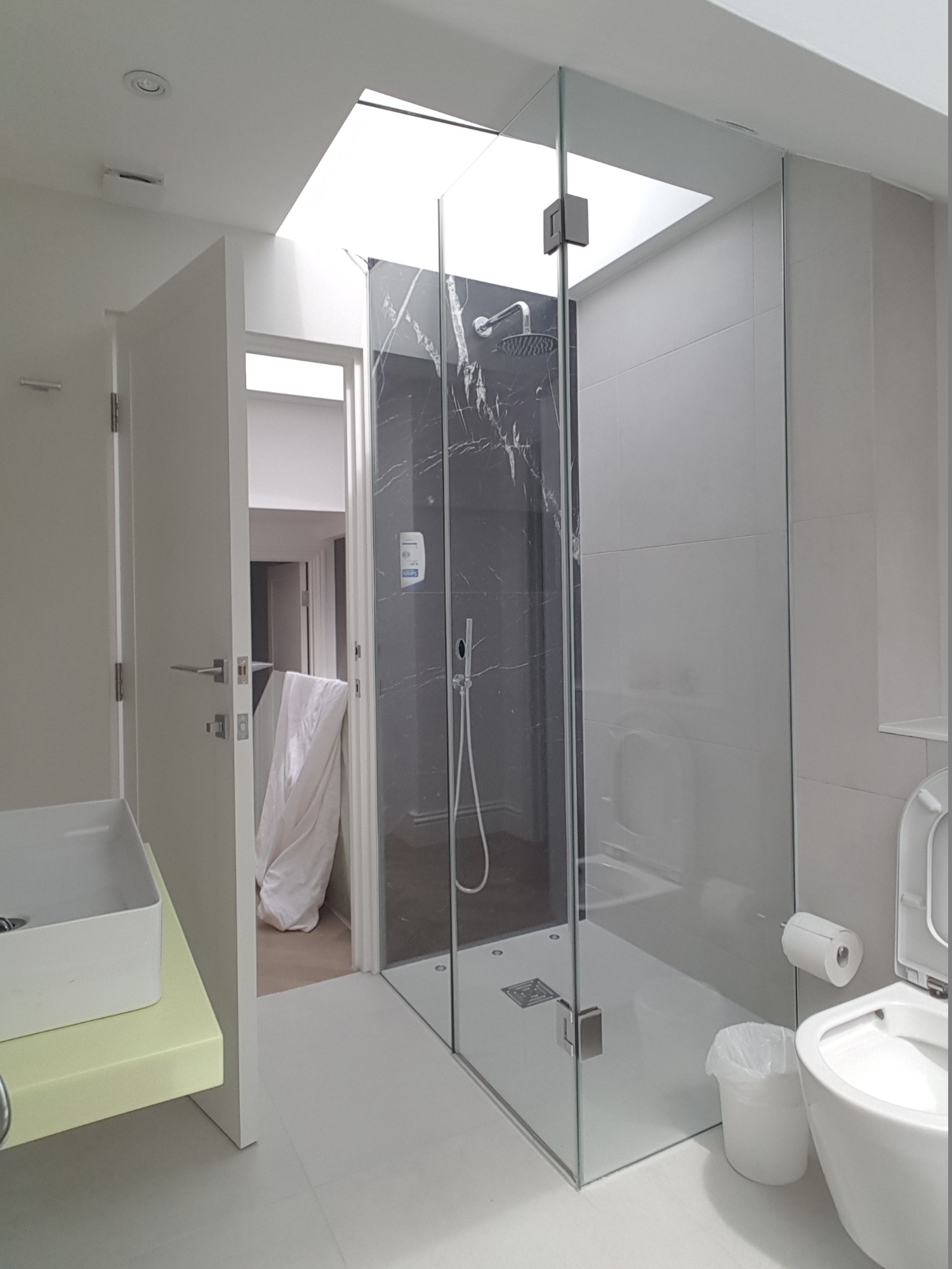 Nowoczesna kabina prysznicowa ze szklanymi ścianami i drzwiami, z czarnym panelem imitującym marmur wewnątrz, oświetlona naturalnym światłem wpadającym przez okno dachowe.