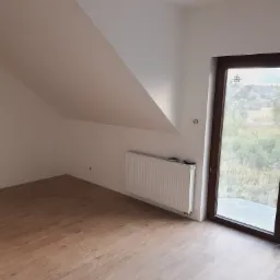 Poddasze po remoncie: panele podłogowe imitujące drewno, białe ściany ze skosem, grzejnik i duże okno z widokiem na zieleń.