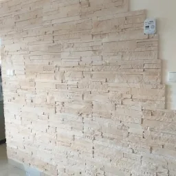 Ściana wyłożona dekoracyjnym, beżowym kamieniem w nieregularnym wzorze, z widocznym czujnikiem dymu na górze i fragmentem podłogi z jasnych płytek