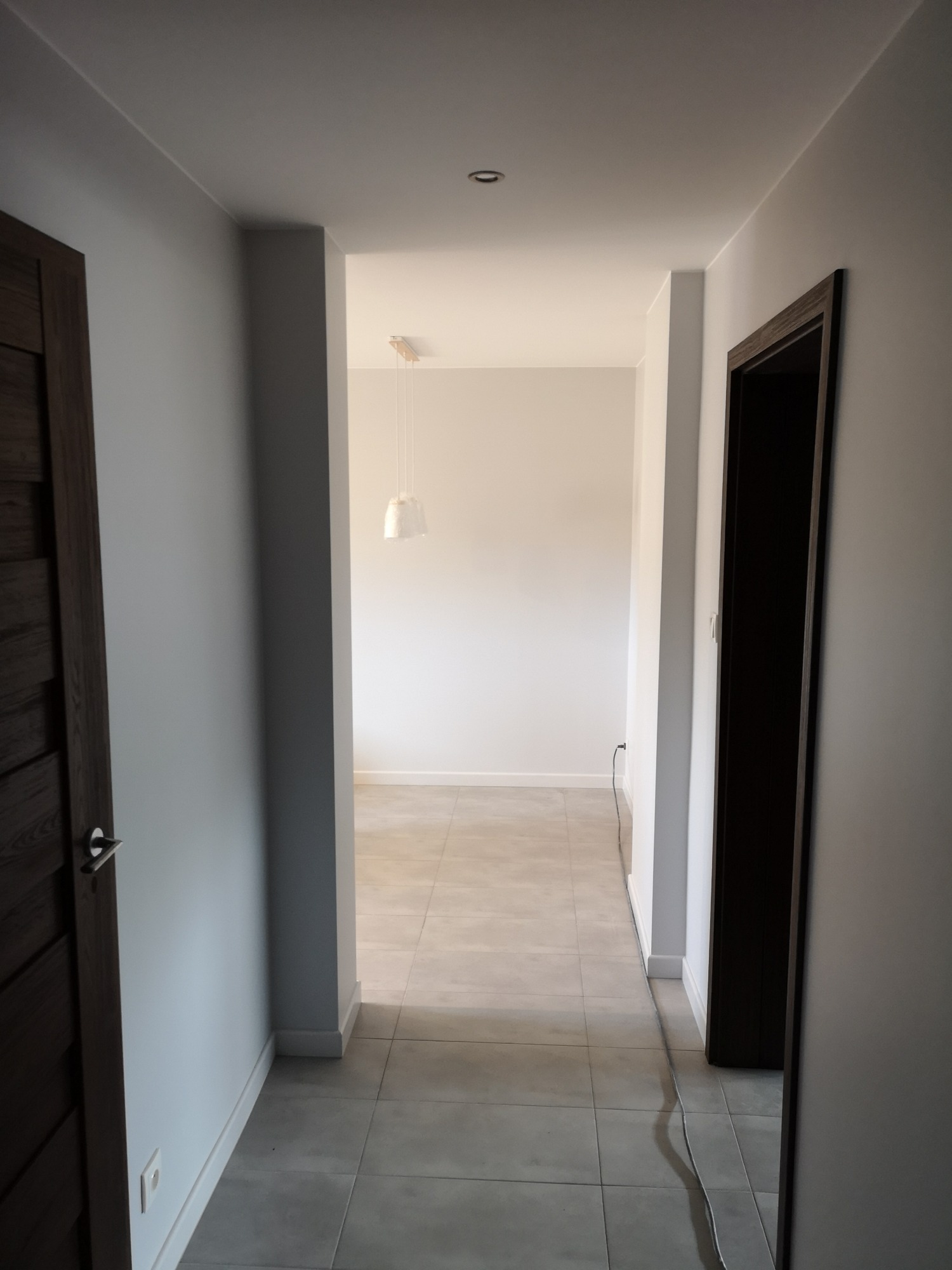 Korytarz w odcieniach szarości z drewnianymi drzwiami, widok na pokój z wiszącą lampą i szarą podłogą, kabel elektryczny wzdłuż ściany.