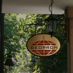 Owalny szyld firmy GEOPRON z logo w formie czerwonej kuli ziemskiej, zawieszony na ozdobnym, kutym wsporniku z latarnią, na tle zieleni drzew