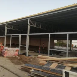 Konstrukcja stalowa hali w trakcie budowy, widoczne słupy i belki, w tle cegły i worki z materiałami budowlanymi, na pierwszym planie deski i gruz.