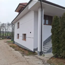 Nowoczesny dom jednorodzinny z białą elewacją, brązowymi oknami i schodami wejściowymi z szarej kostki, otoczony zielenią i drzewami.