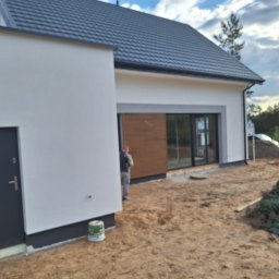 Jacek MALSKI - Nowoczesny dom jednorodzinny z białymi ścianami, drewnianą okładziną, szarą stolarką okienną i ciemnoszarą dachówką, widoczny z zewnątrz w trakcie prac wykończeniowych, z piaskiem na podwórku...