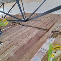 Jacek MALSKI - Montaż konstrukcji stalowej pod sufitem, widoczne drewniane deski, materiały izolacyjne i płyty sufitowe w trakcie prac wykończeniowych.