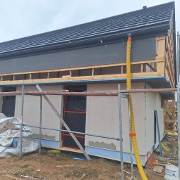 Jacek MALSKI - Budowa domu jednorodzinnego w trakcie prac elewacyjnych i ocieplania, widoczne rusztowania, worki z materiałem izolacyjnym, szara elewacja na parterze, drewniana konstrukcja poddasza z czarną...