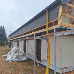 Jacek MALSKI - Budynek w trakcie ocieplania, widoczne rusztowania, drewniana konstrukcja dachu, szare płyty izolacyjne i worki z materiałem budowlanym na ziemi.