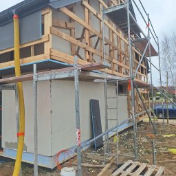 Jacek MALSKI - Budowa domu jednorodzinnego w trakcie prac elewacyjnych z widocznym rusztowaniem, drewnianą konstrukcją szkieletową, żółtą rurą wentylacyjną i częściowo otynkowaną ścianą zewnętrzną.