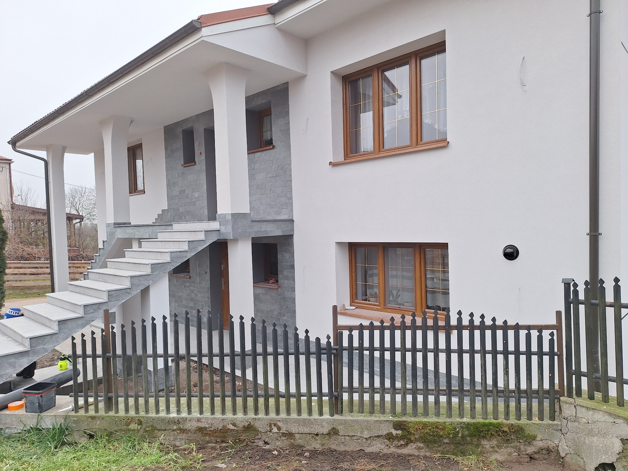 Dwupiętrowy dom z białą elewacją, brązowymi oknami, szarymi elementami dekoracyjnymi i czarnym ogrodzeniem, widok z zewnątrz w pochmurny dzień.