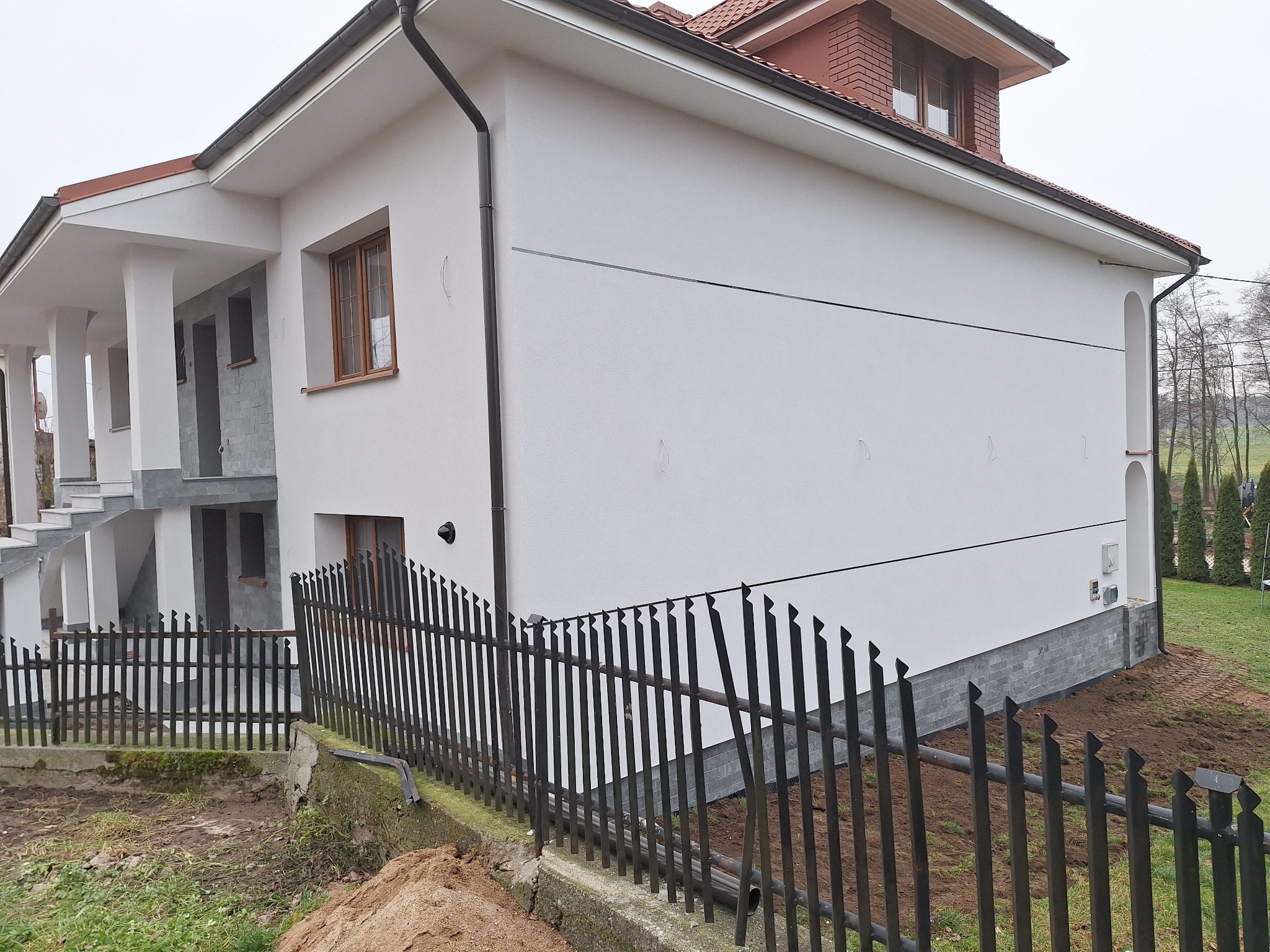 Nowoczesny dom z białą elewacją, widoczne fragmenty czarnego ogrodzenia, szara podmurówka i okna z drewnianymi ramami.
