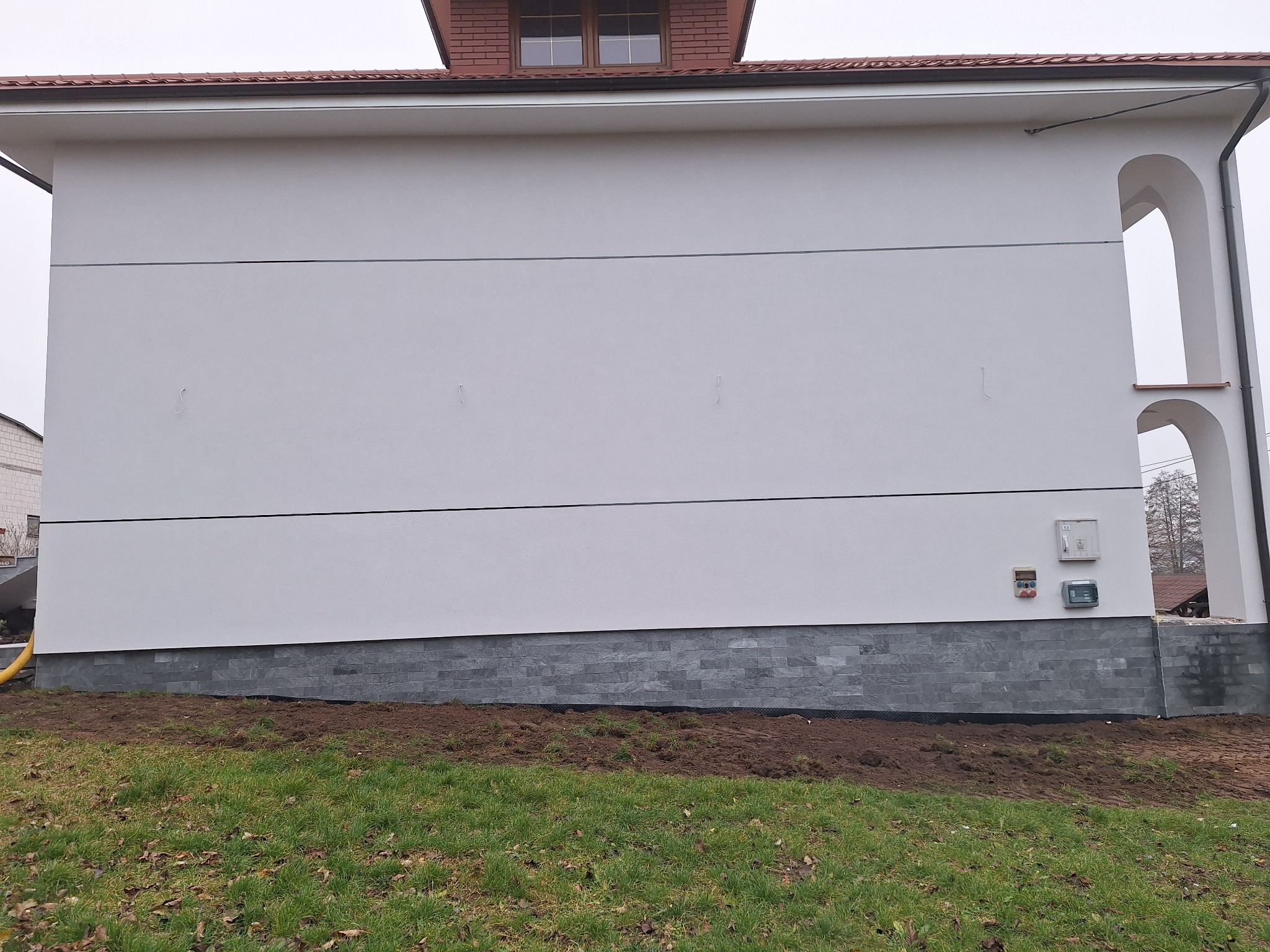 Świeżo otynkowana, biała elewacja budynku z szarą, kamienną podmurówką i widocznymi skrzynkami elektrycznymi, otoczona trawnikiem i przygotowaną ziemią pod nasadzenia.