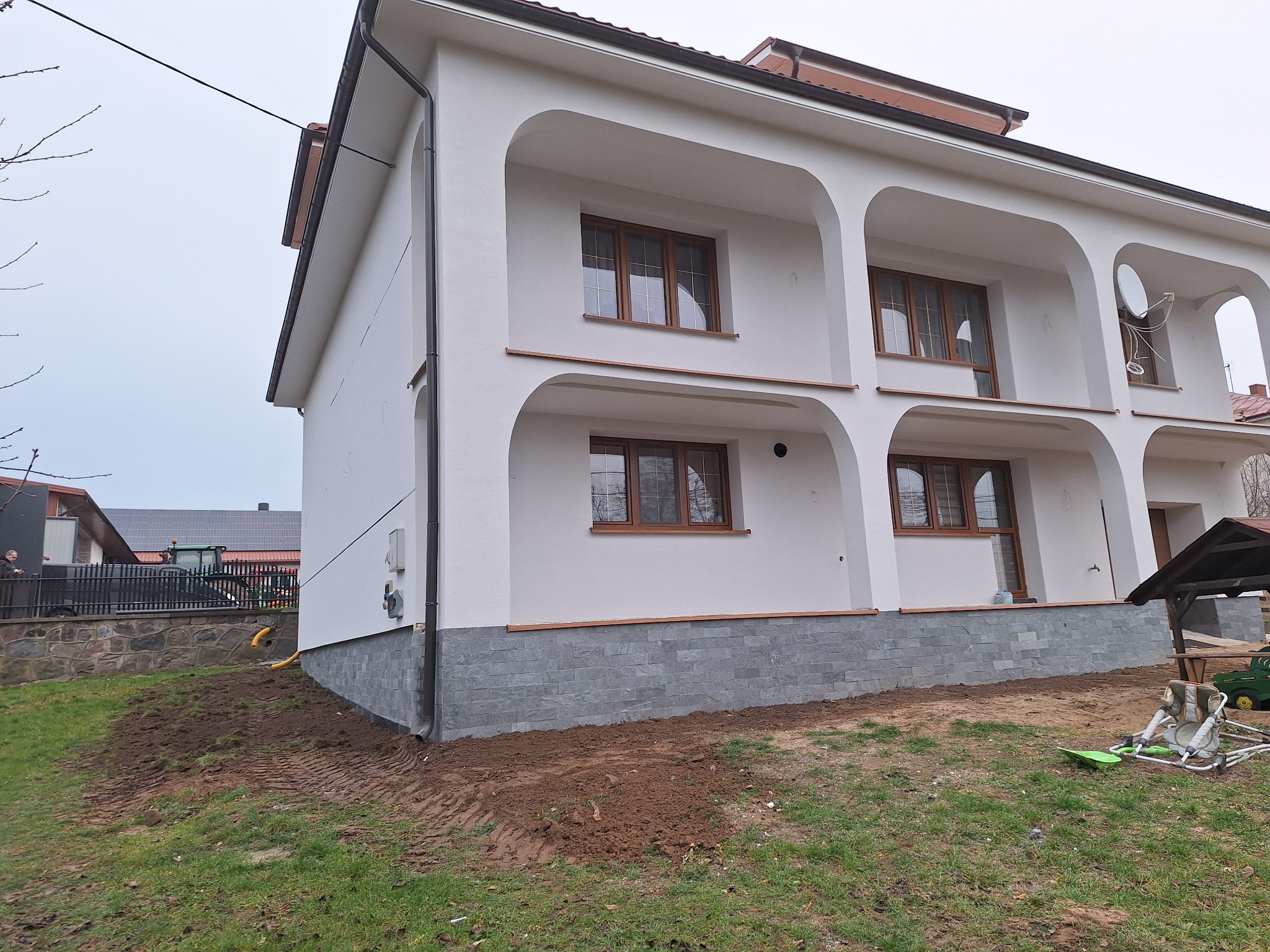 Dwupiętrowy dom z białymi ścianami, balkonami z łukami i szarą podmurówką z kamienia, widok z boku na tle pochmurnego nieba.