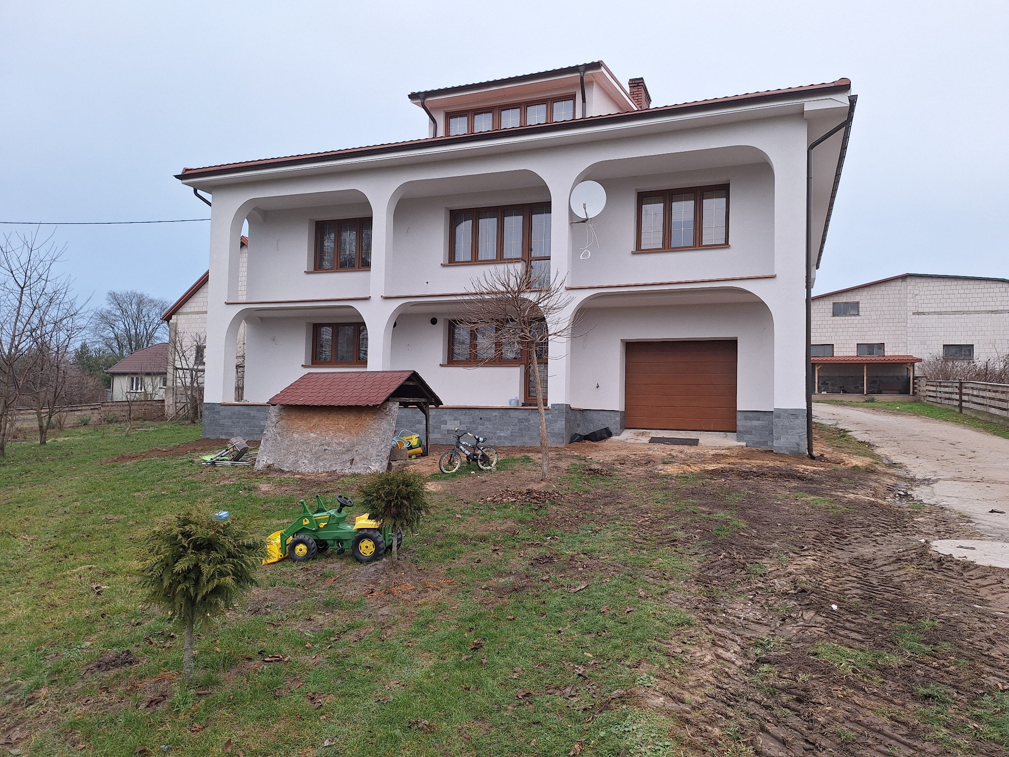 Dwupiętrowy dom z białymi ścianami i brązowymi oknami, widoczna antena satelitarna, na trawniku zabawkowy traktor i rower.
