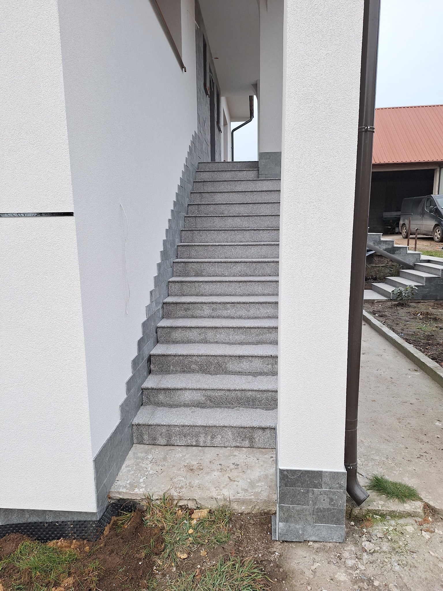 Zewnętrzne schody wejściowe wykończone szarą kostką brukową, prowadzące do budynku z białą, teksturowaną elewacją i szarym cokołem.