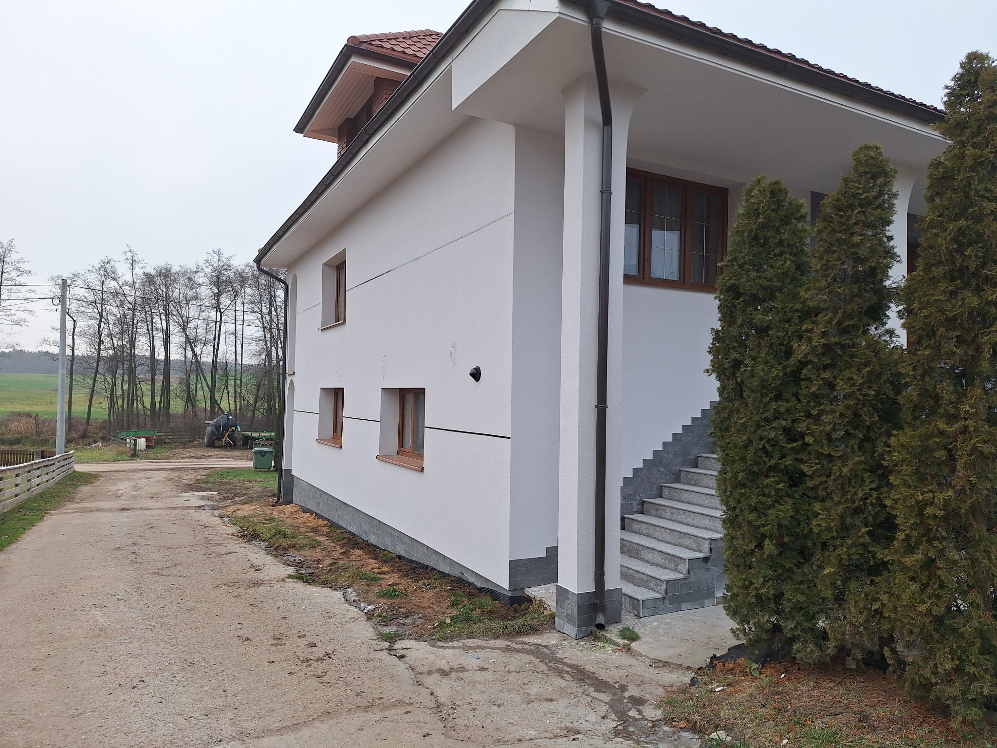 Nowoczesny dom jednorodzinny z białą elewacją, brązowymi oknami i schodami wejściowymi z szarej kostki, otoczony zielenią i drzewami.