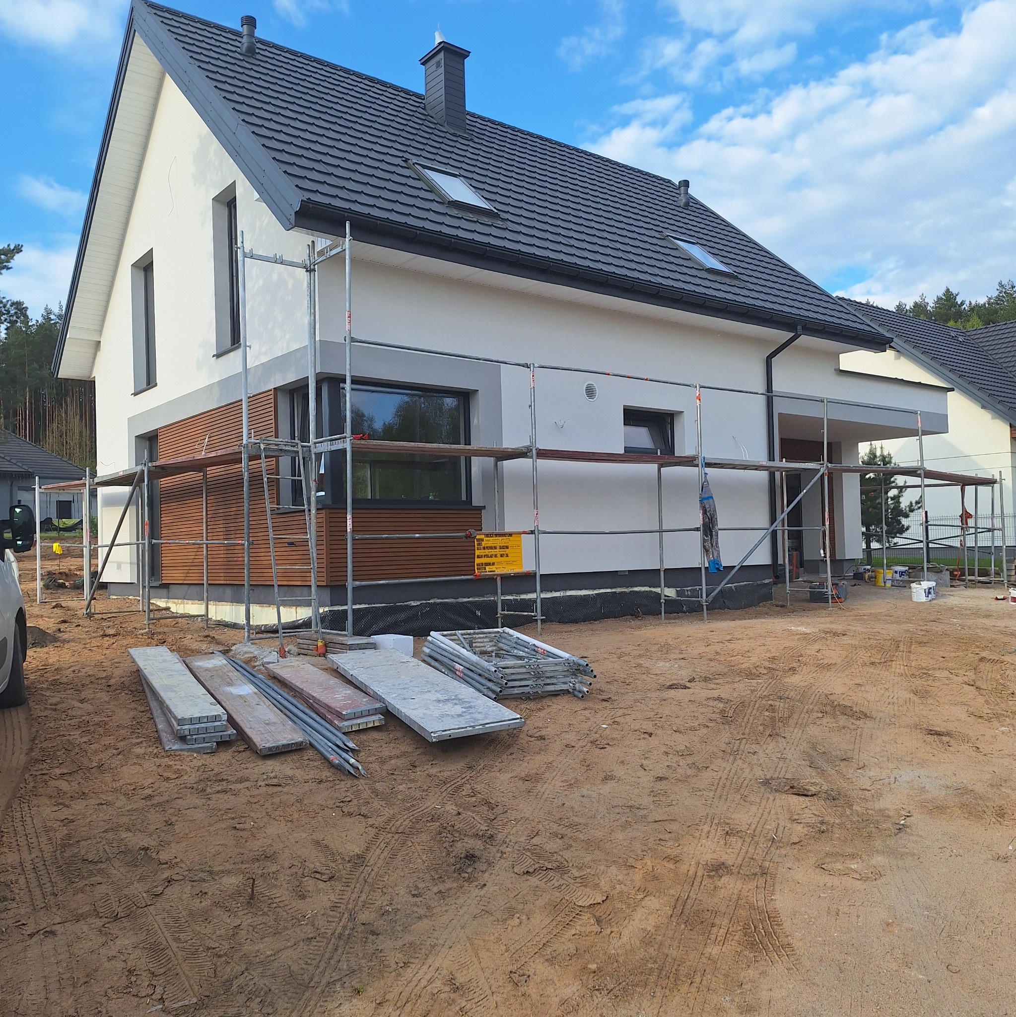 Dom jednorodzinny w trakcie prac wykończeniowych elewacji, widoczne rusztowania, deski elewacyjne oraz przygotowany teren wokół budynku.