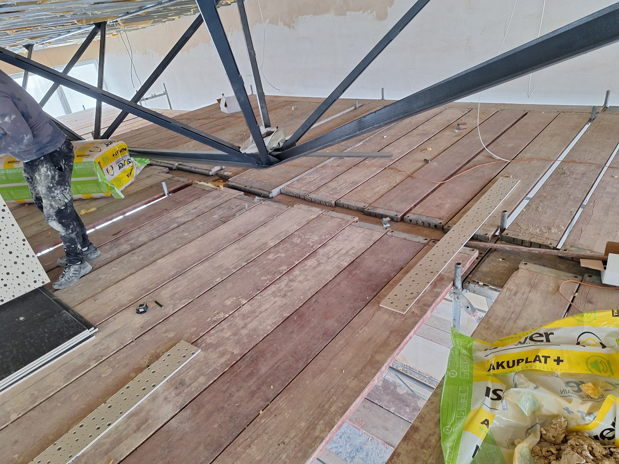 Montaż konstrukcji stalowej pod sufitem, widoczne drewniane deski, materiały izolacyjne i płyty sufitowe w trakcie prac wykończeniowych.