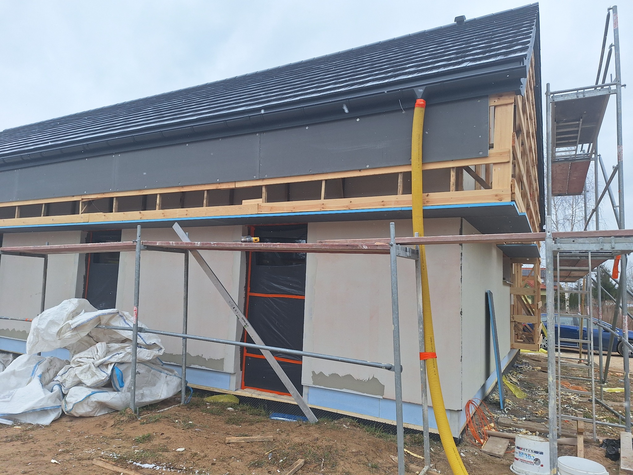 Budowa domu jednorodzinnego w trakcie prac elewacyjnych i ocieplania, widoczne rusztowania, worki z materiałem izolacyjnym, szara elewacja na parterze, drewniana konstrukcja poddasza z czarną...