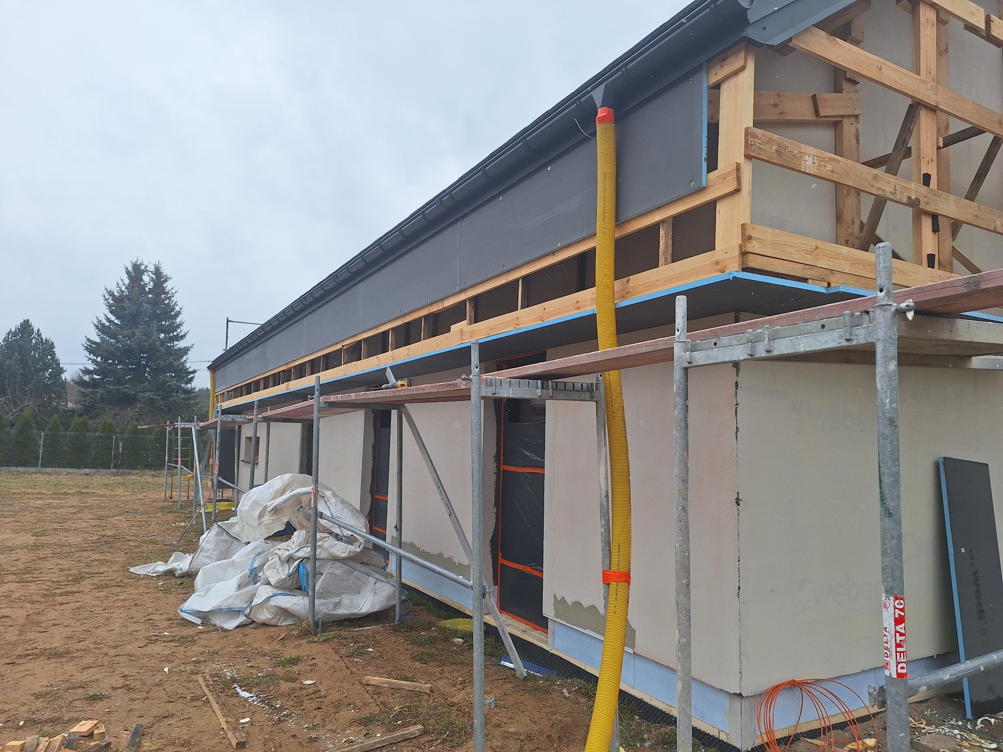 Budynek w trakcie ocieplania, widoczne rusztowania, drewniana konstrukcja dachu, szare płyty izolacyjne i worki z materiałem budowlanym na ziemi.