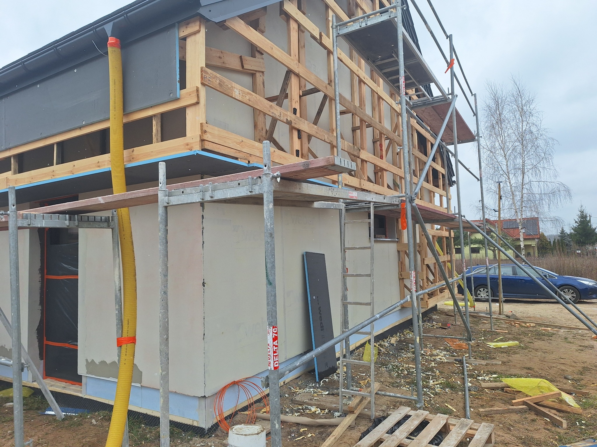Budowa domu jednorodzinnego w trakcie prac elewacyjnych z widocznym rusztowaniem, drewnianą konstrukcją szkieletową, żółtą rurą wentylacyjną i częściowo otynkowaną ścianą zewnętrzną.