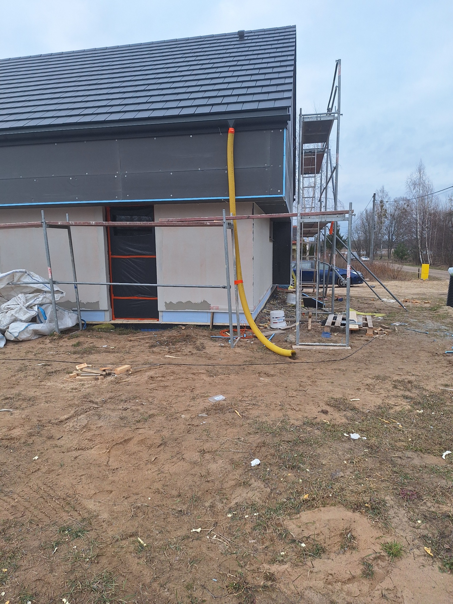 Budynek w trakcie ocieplania; widoczne rusztowania, żółty wąż wentylacyjny, czarna membrana na elewacji i częściowo otynkowane ściany.