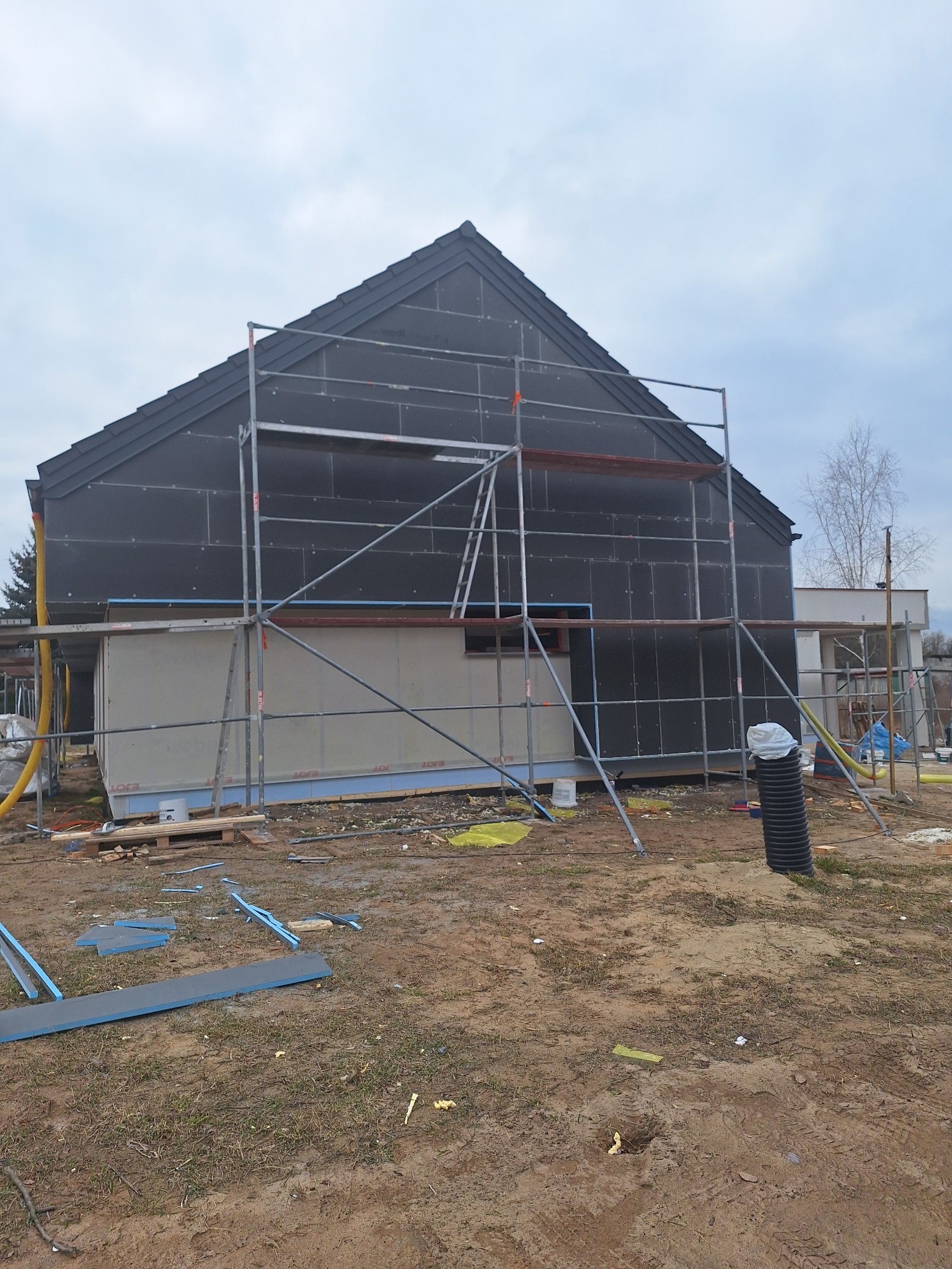 Dom w trakcie ocieplania elewacji, widoczne rusztowanie, czarne płyty izolacyjne na dachu i ścianach, oraz fragment otynkowanej ściany.