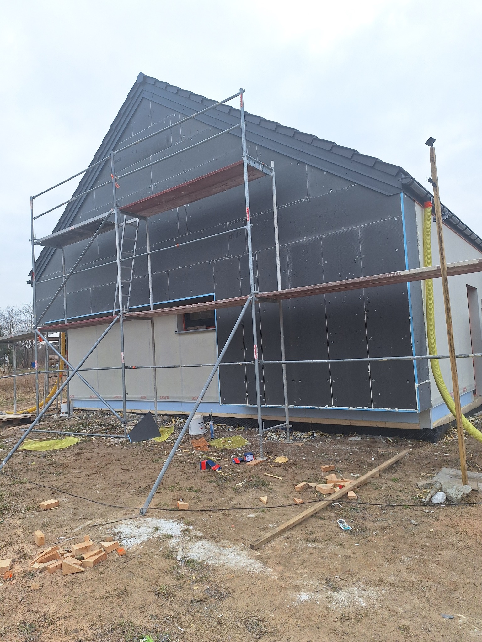 Dom w trakcie ocieplania, widoczne rusztowania, czarne płyty izolacyjne na ścianach, żółty wąż wentylacyjny i porozrzucane materiały budowlane na ziemi.