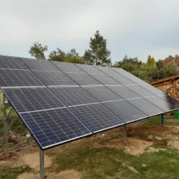 Panel słoneczny na metalowej konstrukcji ustawiony na trawie obok składu drewna, w tle drzewa i dom.