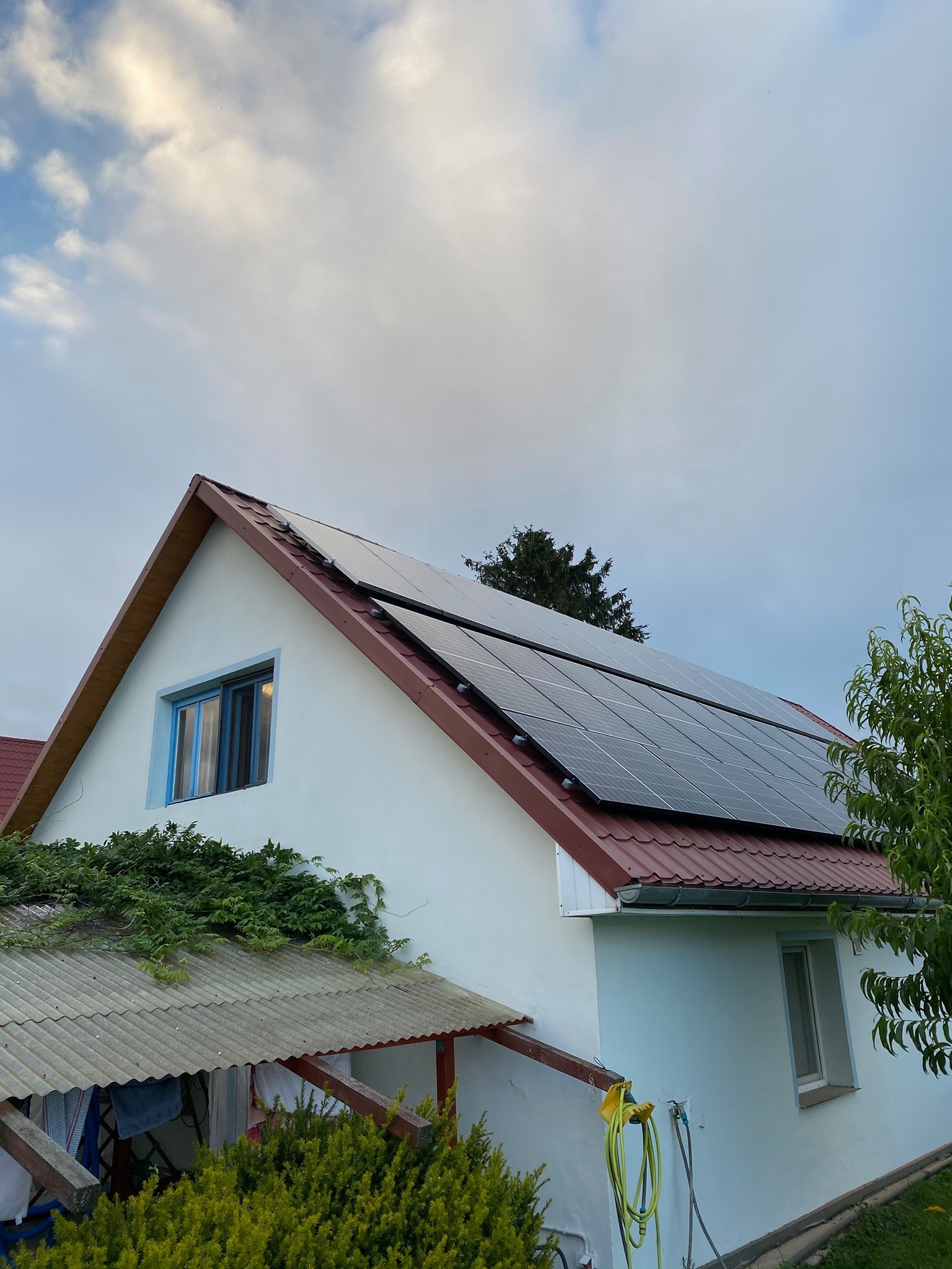 Dom jednorodzinny z panelami fotowoltaicznymi na dachu pokrytym czerwoną dachówką, widok z dołu na tle pochmurnego nieba.