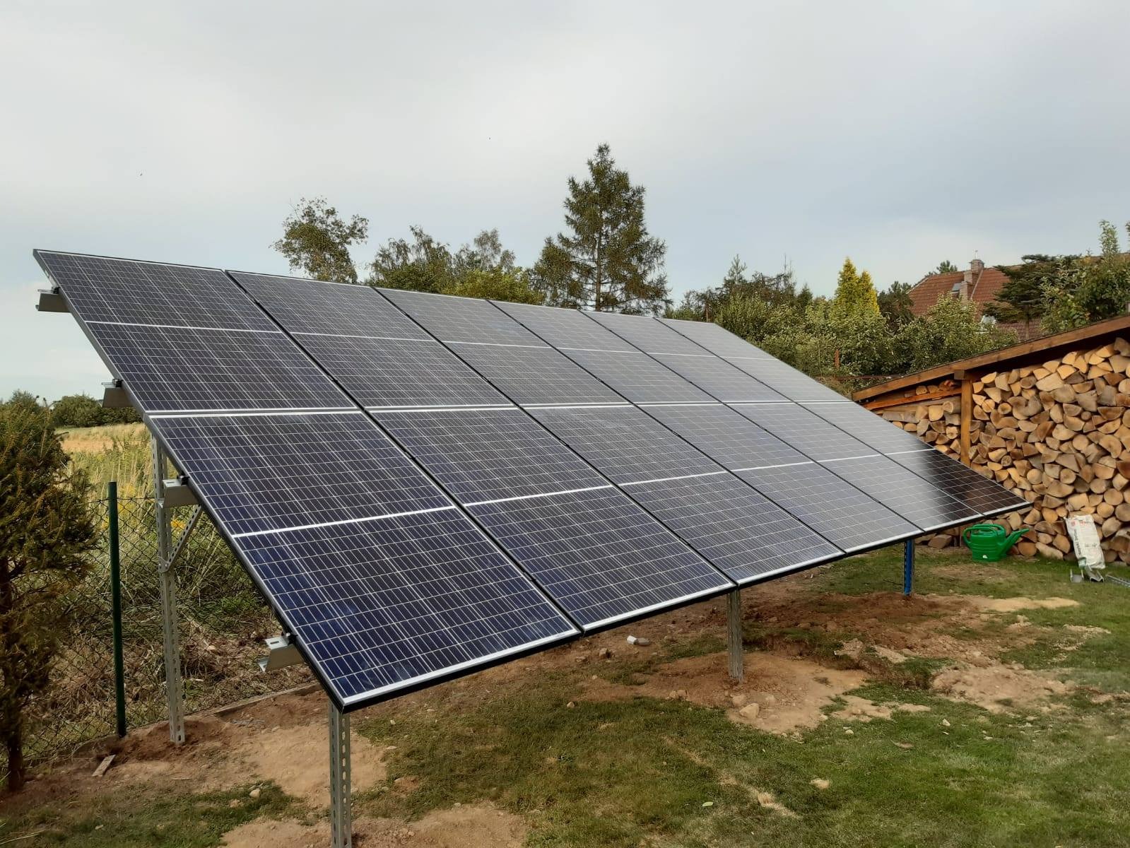 Panel słoneczny na metalowej konstrukcji ustawiony na trawie obok składu drewna, w tle drzewa i dom.