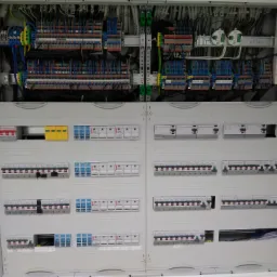 Otwarta szafa elektryczna z widocznymi zabezpieczeniami, przewodami i listwami zaciskowymi wewnątrz, część wyłączników jest wyłączona.