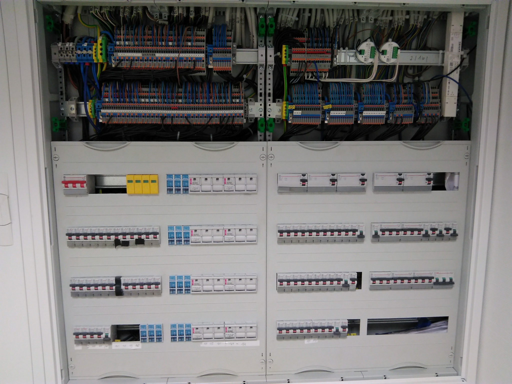 Otwarta szafa elektryczna z widocznymi zabezpieczeniami, przewodami i listwami zaciskowymi wewnątrz, część wyłączników jest wyłączona.