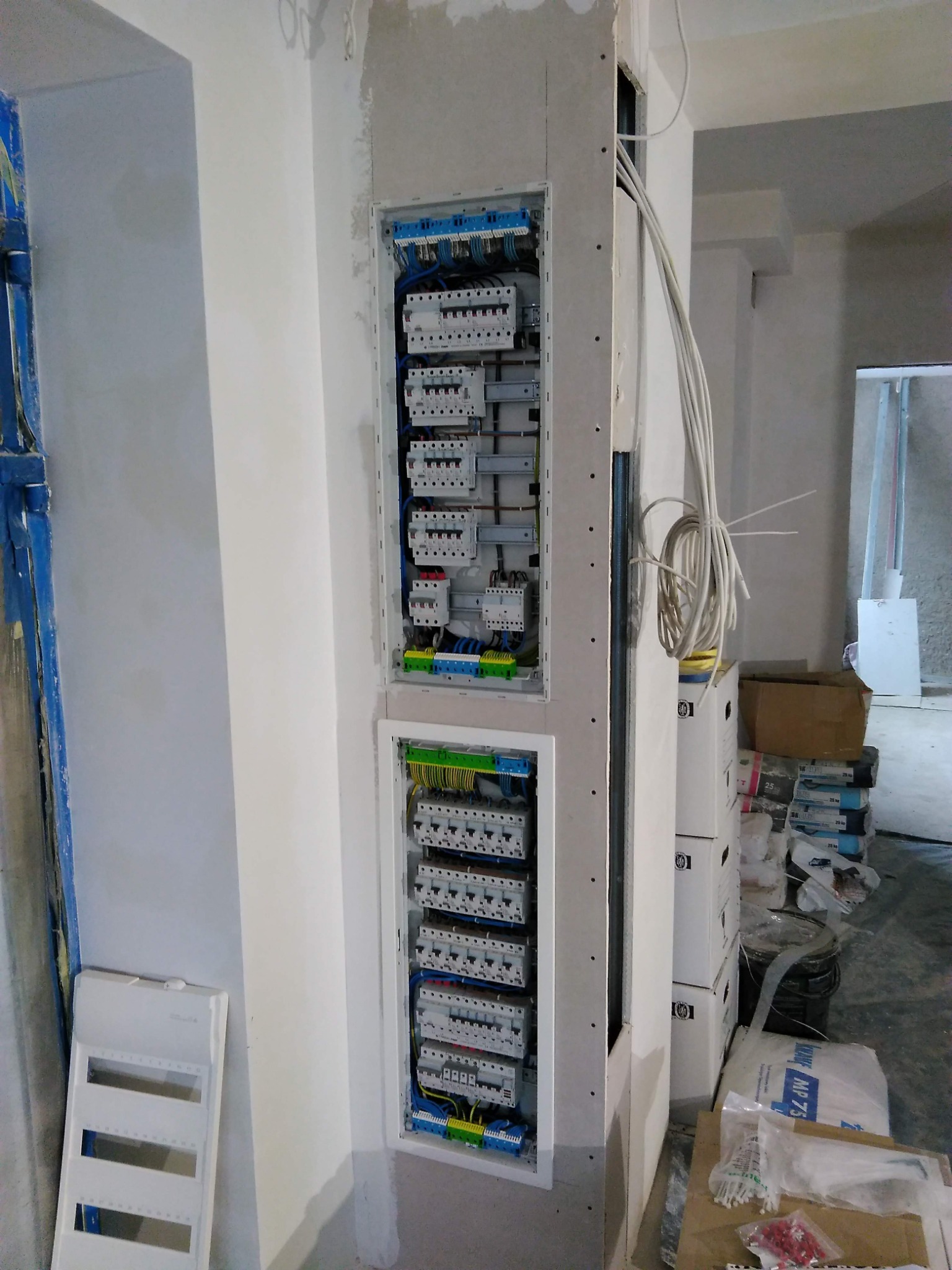 Dwa panele elektryczne z zainstalowanymi bezpiecznikami i okablowaniem, umieszczone w ścianie pokrytej płytą gipsowo-kartonową, widoczne przewody elektryczne i materiały budowlane w tle.