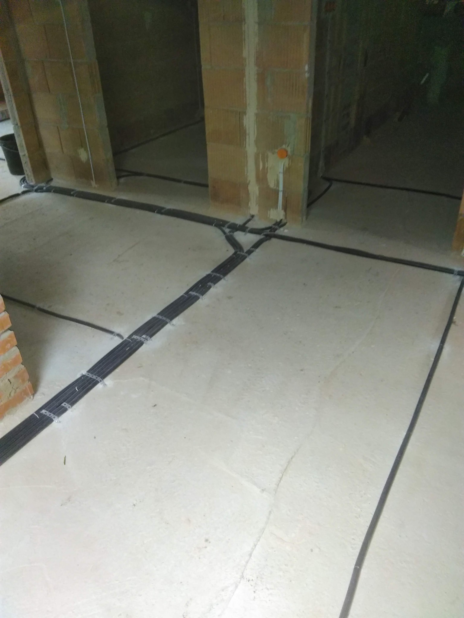 Rozprowadzenie przewodów elektrycznych w peszlach, przymocowanych metalowymi uchwytami do surowej posadzki w budynku w trakcie budowy, widoczne ściany z pustaków.