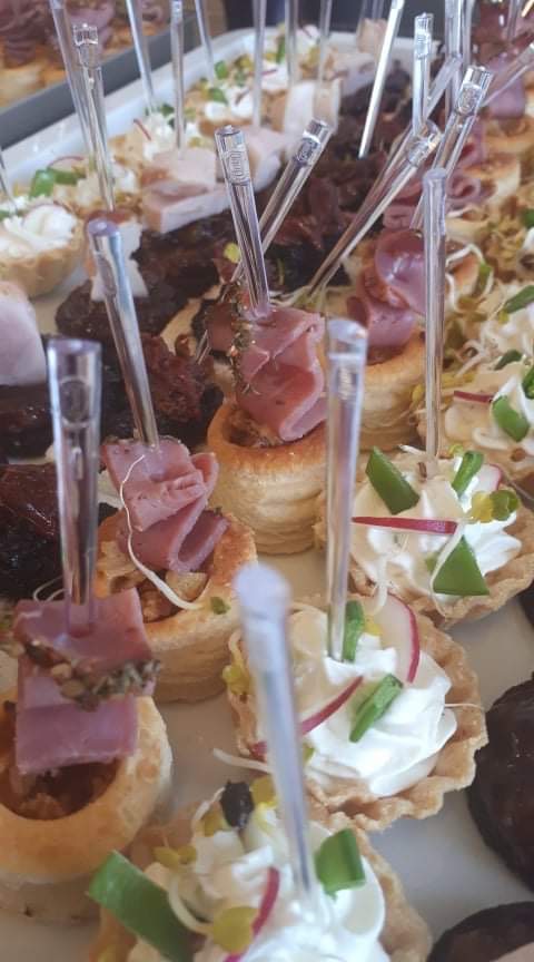 Różnorodne przekąski na tacy, w tym mini tartaletki z kremem i warzywami oraz vol-au-venty z wędliną, spięte przezroczystymi wykałaczkami, ułożone blisko siebie.